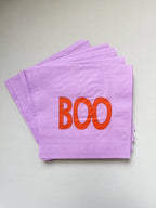A stack of light purple paper napkins with the word 'BOO' printed in orange across the top napkin, photographed on a plain light gray surface.