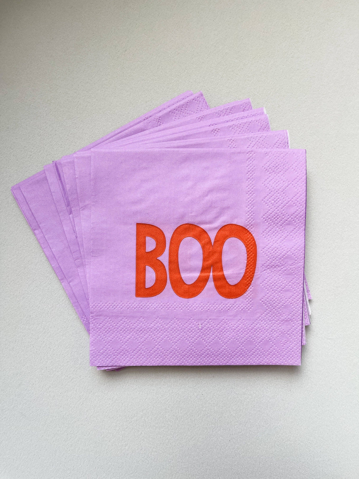 A stack of light purple paper napkins with the word 'BOO' printed in orange across the top napkin, photographed on a plain light gray surface.