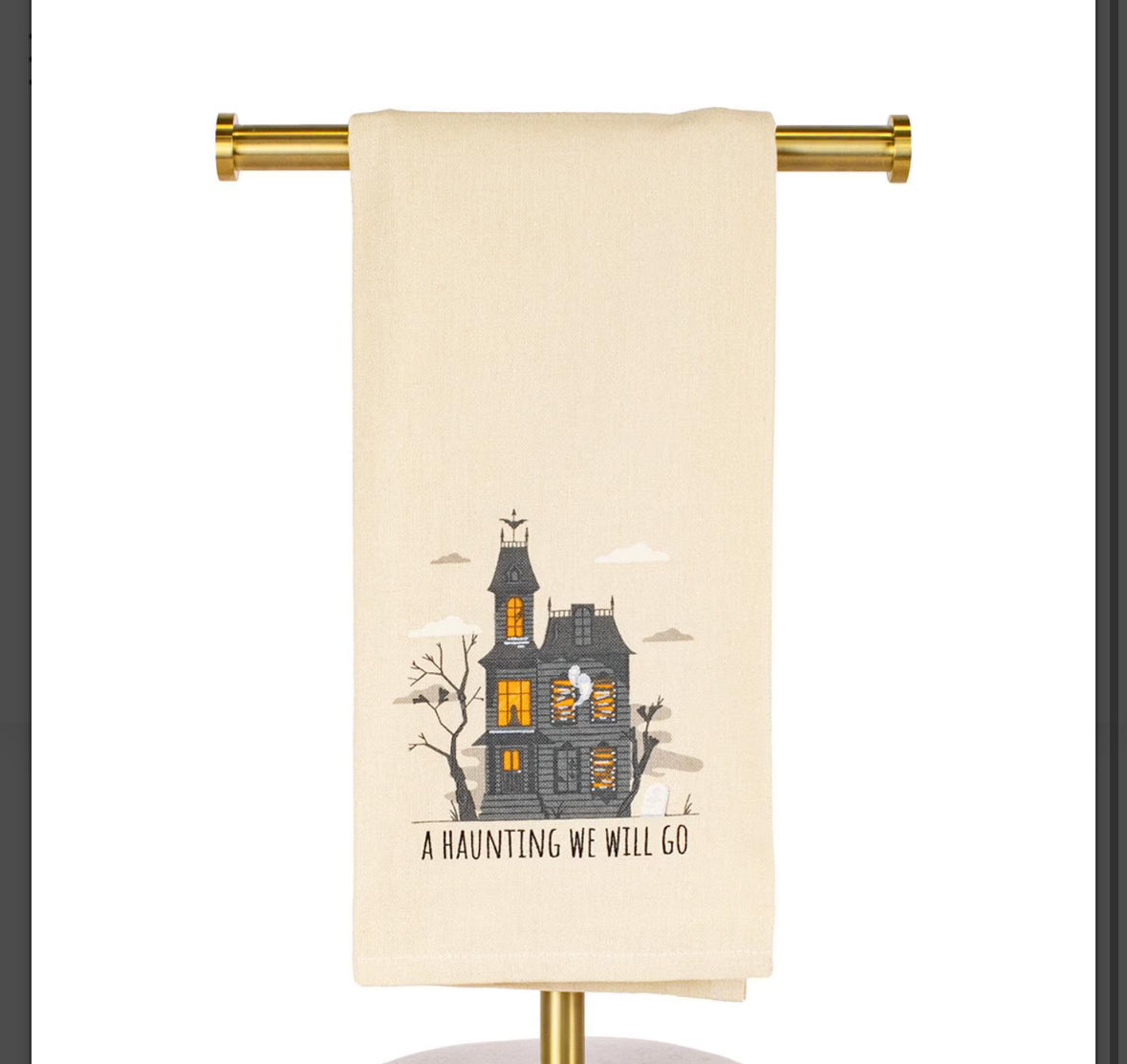 A cream-colored kitchen towel with a Halloween graphic of a haunted house and the text 'A HAUNTING WE WILL GO', hanging from a gold metal rod against a white background.