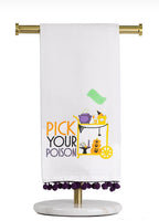 A white kitchen towel with purple pom-pom trim hanging from a gold stand. The towel features a colorful graphic of a yellow cat in a witch hat next to a serving cart with pumpkins, with the text 'PICK YOUR POISON' in orange and purple.