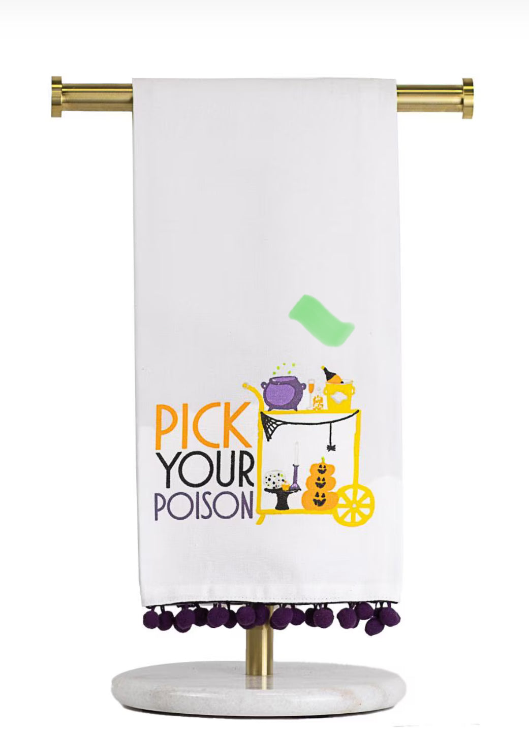 A white kitchen towel with purple pom-pom trim hanging from a gold stand. The towel features a colorful graphic of a yellow cat in a witch hat next to a serving cart with pumpkins, with the text 'PICK YOUR POISON' in orange and purple.
