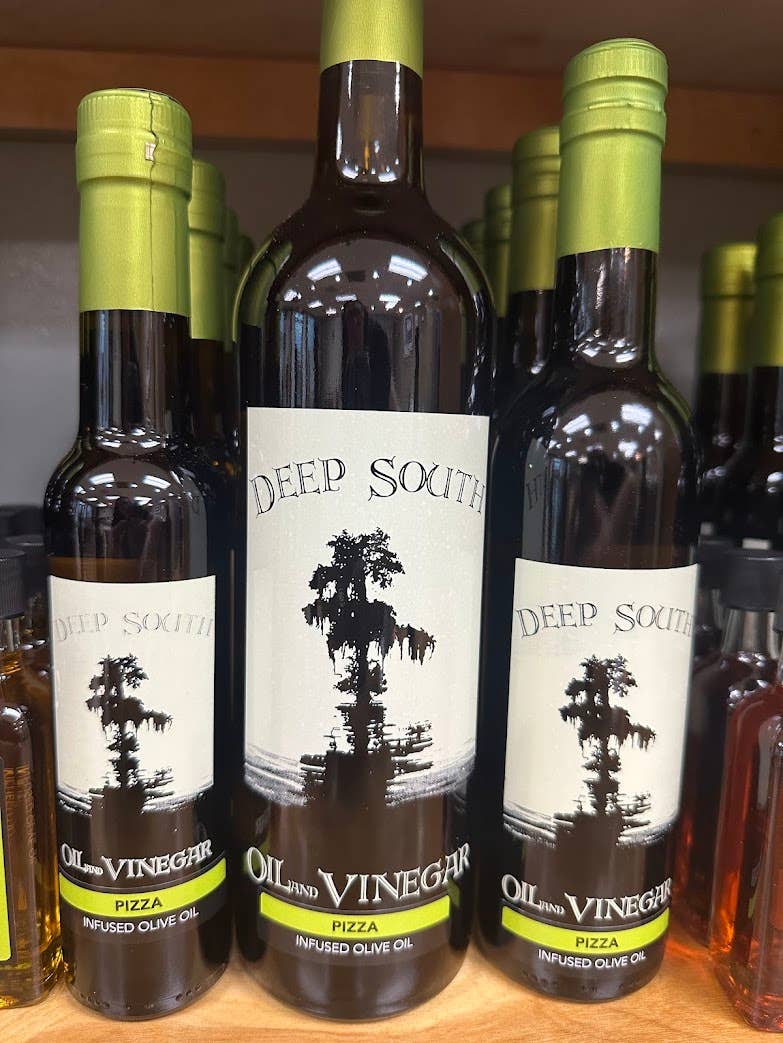 Three bottles of Deep South Oil and Vinegar Pizza Infused Olive Oil displayed on a wooden shelf, with the center bottle featured prominently and other bottles blurred in the background.