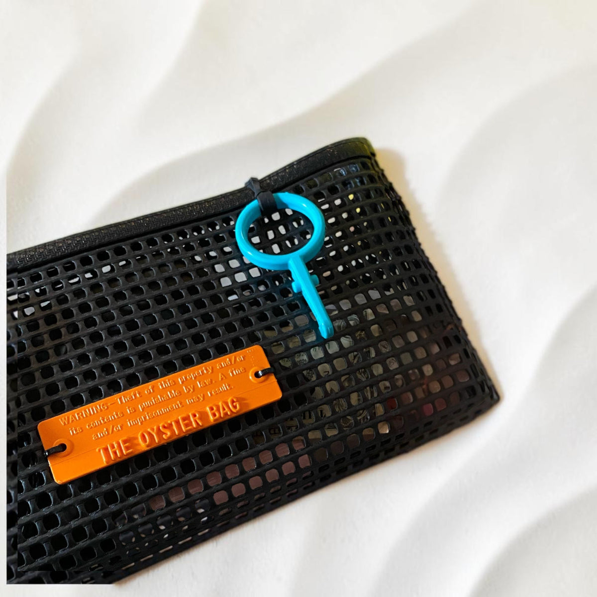 FULL MOOD black mesh clutch bag with an orange branded tag and blue key ring on a marble table next to drinks