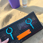 FULL MOOD The Clutch oyster bag in black mesh with teal rings and orange label on a sandy beach