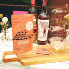 FULL MOOD Stir Sessions blood orange old fashioned cocktail mix packets on a wooden board with bourbon and a glass.