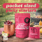 FULL MOOD Stir Sessions Cranberry Mule cocktail mix packets with a prepared drink in a copper mug on a picnic table.