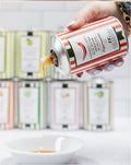 FULL MOOD hand pouring spicy Peperoncino extra virgin olive oil from a decorative striped tin into a white bowl.