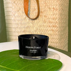 FULL MOOD Hidden Forest soy wax candle in a black glass jar with an embedded gemstone, resting on a large green leaf.