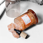 An amber glass jar tipped on its side with pink Himalayan salt spilling out onto a white surface, next to a small black dropper bottle labeled Ginko Oil and an empty clear glass jar in the background.