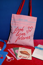 FULL MOOD pink canvas tote bag with library card print and red straps, displayed with books on a red surface.