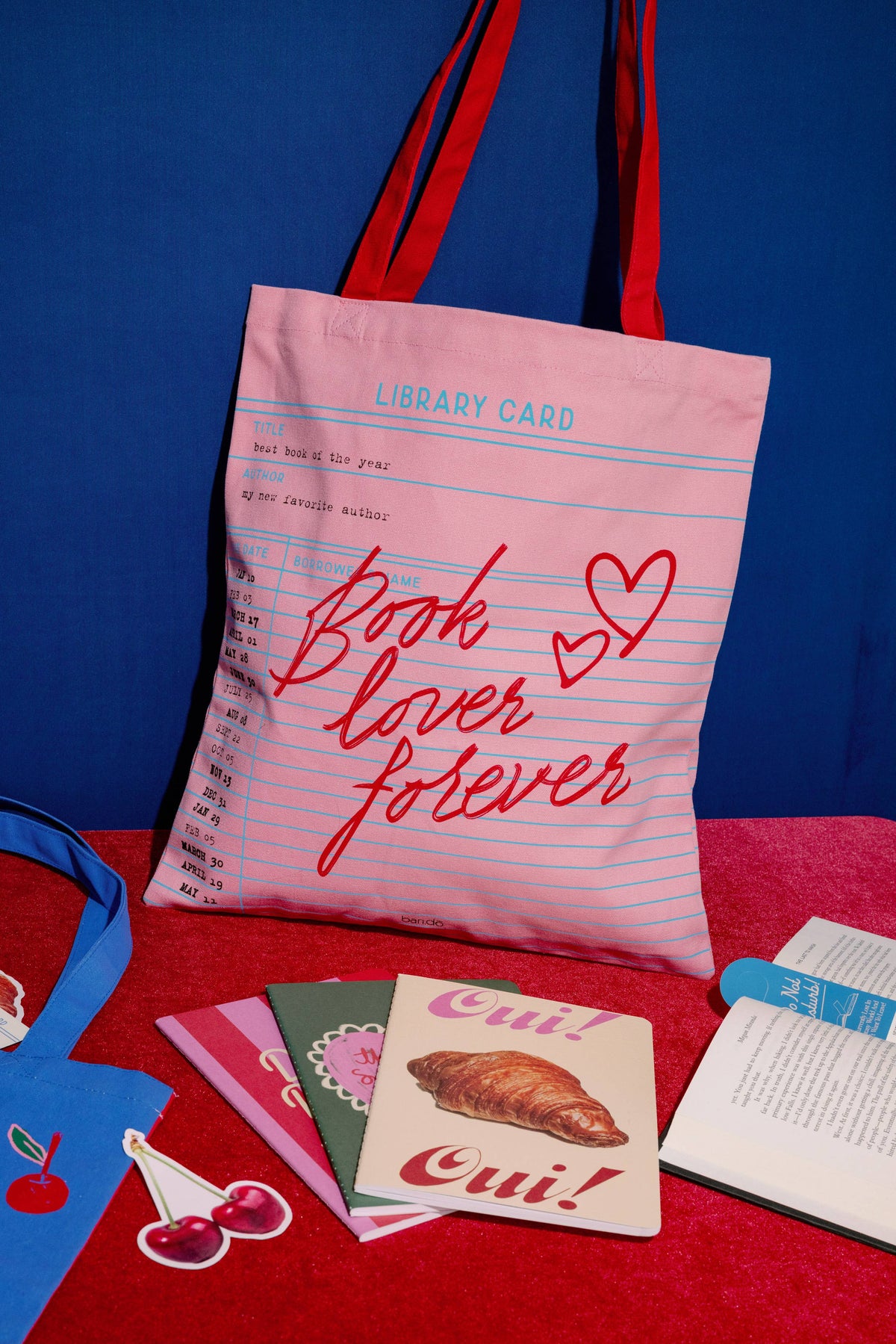 FULL MOOD pink canvas tote bag with library card print and red straps, displayed with books on a red surface.