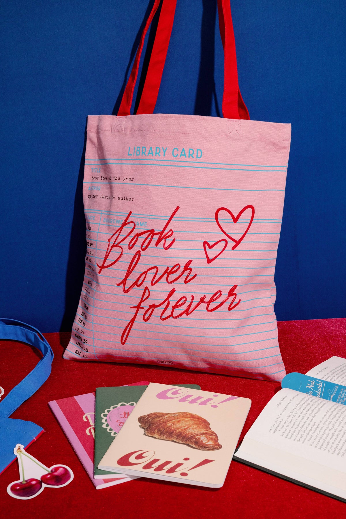 FULL MOOD pink canvas tote bag with library card print and red straps, displayed with notebooks on a red surface.