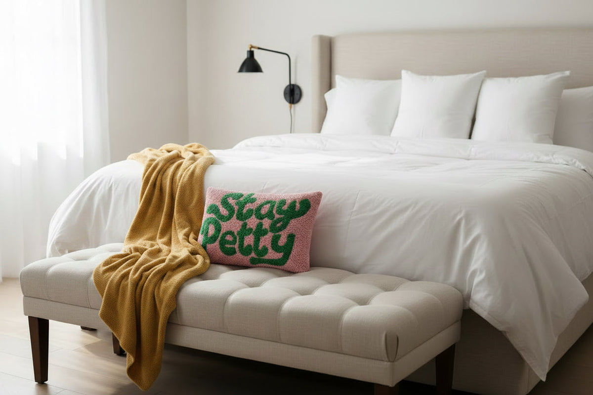 FULL MOOD Stay Petty hook pillow with green text on pink wool, styled on a bedroom bench with a yellow throw blanket.
