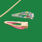 Two decorative hair claw clips on a textured gray surface. The top clip is mint green with colorful rhinestones, and the bottom clip has a pink inner band and gold-tone metal frame.