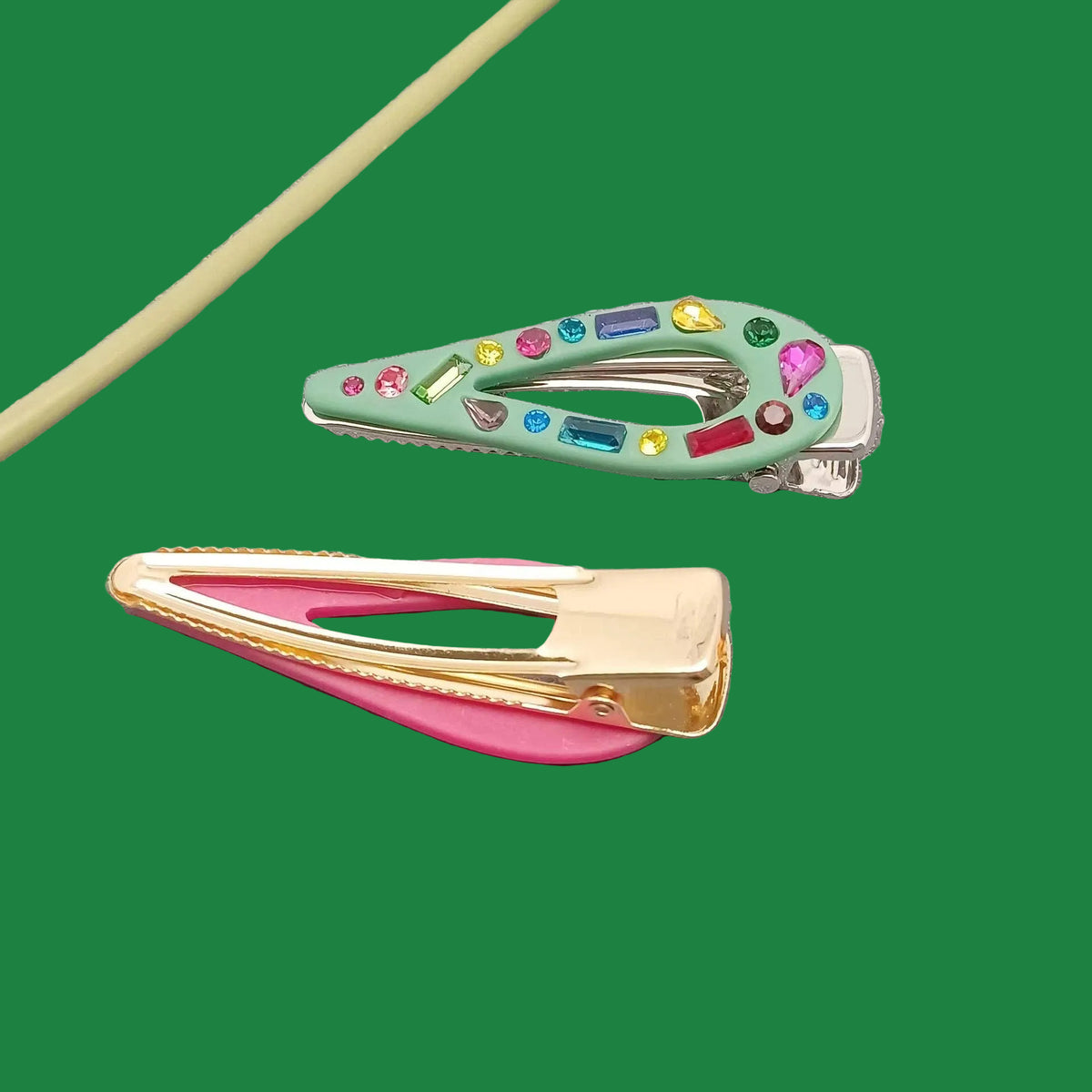 Two decorative hair claw clips on a textured gray surface. The top clip is mint green with colorful rhinestones, and the bottom clip has a pink inner band and gold-tone metal frame.