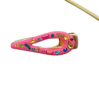 A pink hair clip decorated with colorful faux rhinestones, resting on a textured gray surface with green leaves and a yellow stick in the background.