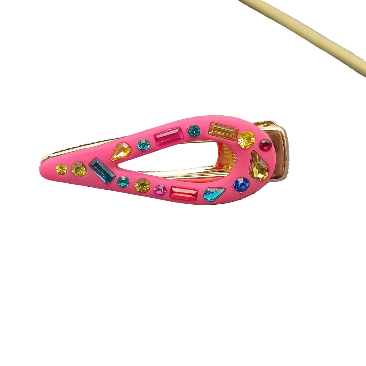 A pink hair clip decorated with colorful faux rhinestones, resting on a textured gray surface with green leaves and a yellow stick in the background.