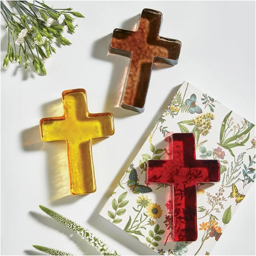 Three translucent cross-shaped soaps in amber, dark brown, and deep red, styled on a white background with a floral-patterned card and green botanical props.