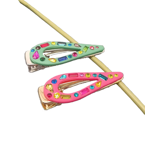 Two hair clips on a textured gray surface, one mint green and one pink, both decorated with colorful faux rhinestones and shown with a gold-tone metal clip mechanism.