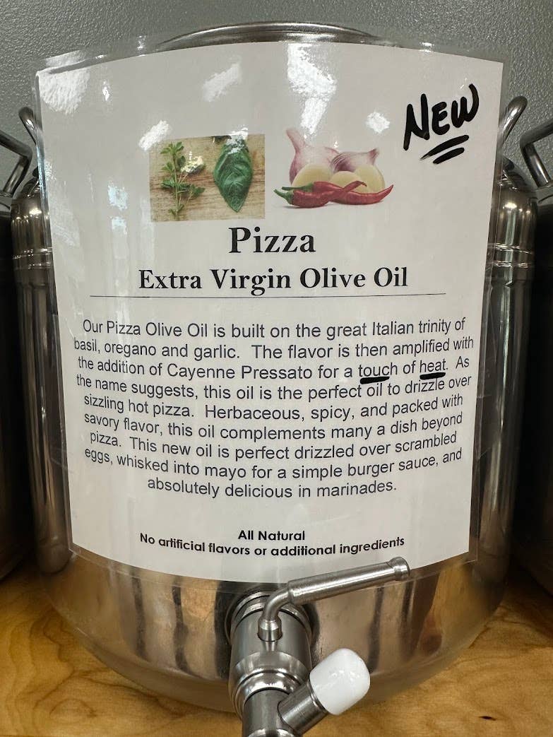 A stainless steel dispenser bottle of Pizza Extra Virgin Olive Oil with a large white product label showing ingredients and descriptive text, set on a wooden surface.
