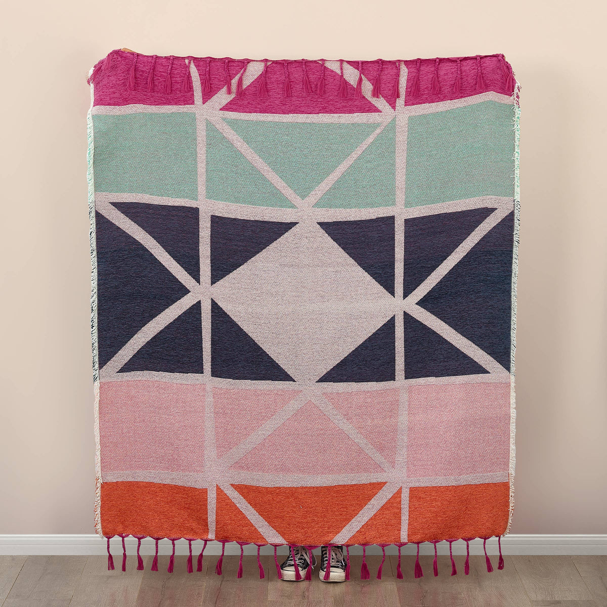 A woven geometric throw blanket with fringe, featuring a multicolor pattern of pink, mint green, navy blue, light gray, and orange blocks, shown hanging against a beige wall with a person's shoes visible at the bottom.