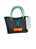 A black plastic straw tote bag with thick mint green rope handles and a turquoise key-shaped charm, displayed on an orange plastic crate at a beach with the ocean and blue sky in the background.