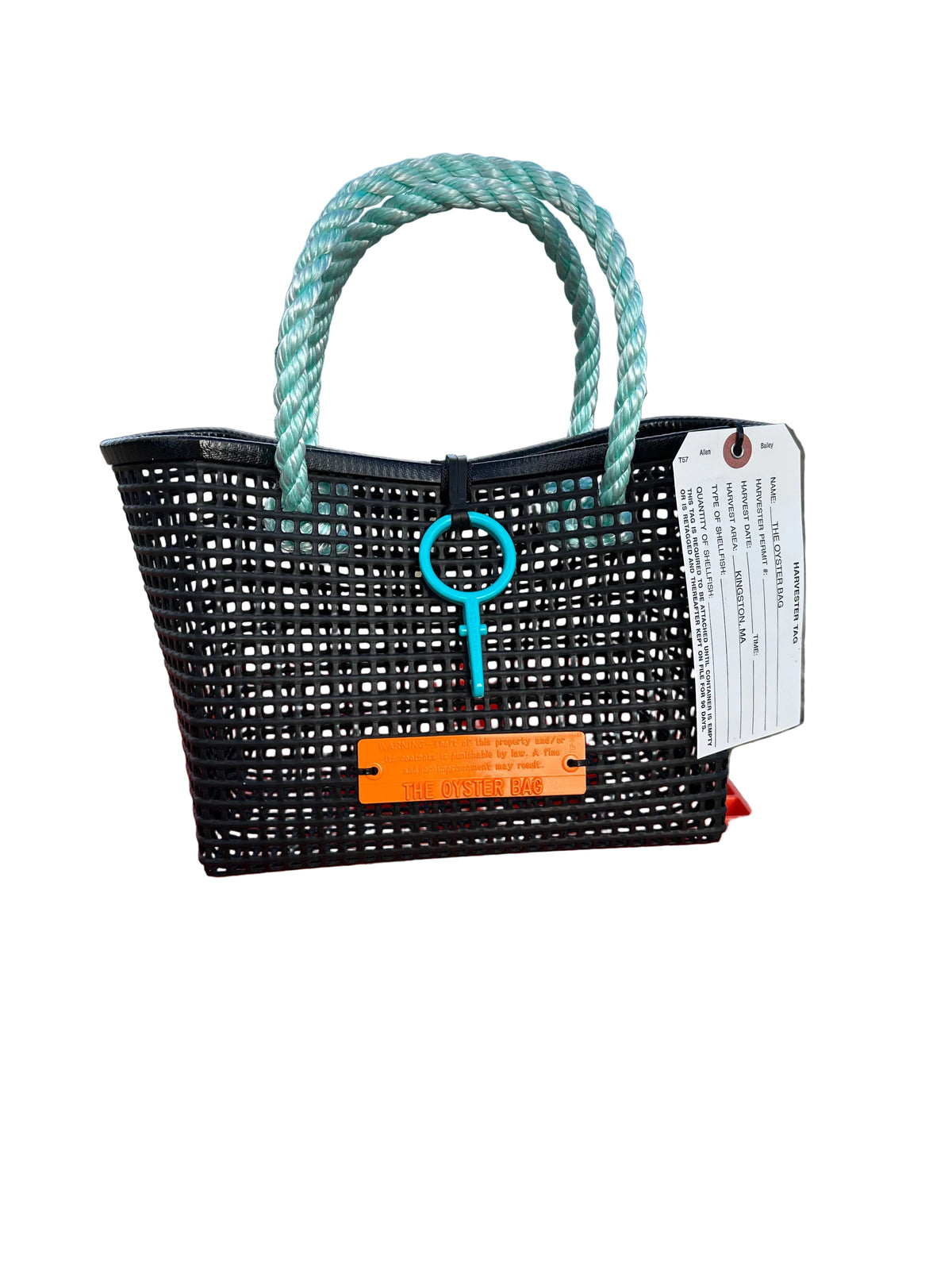 A black plastic straw tote bag with thick mint green rope handles and a turquoise key-shaped charm, displayed on an orange plastic crate at a beach with the ocean and blue sky in the background.